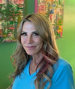 Smiling woman with long blonde hair wearing a blue top against a colorful background.